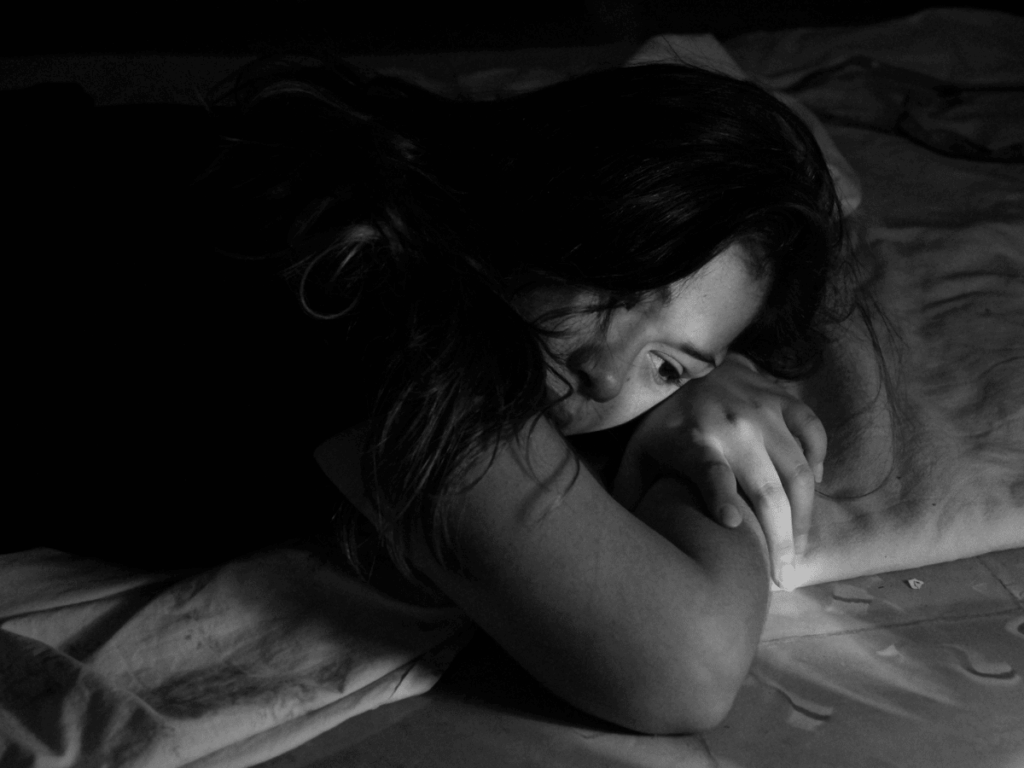 Black and white photo of a woman in bed, lying on her stomach with her eyes open. Sad.