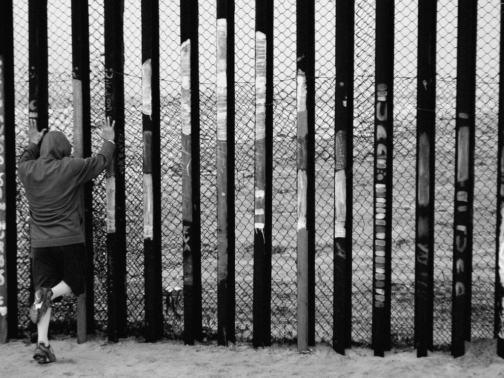 Man with hoody seen from the back, leaning against a rough iron fence.